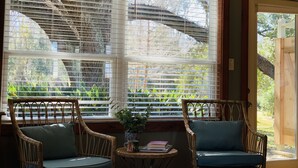 Una habitación con dos sillas de mimbre, una mesita con una planta y libros, y un ventanal con persianas.
