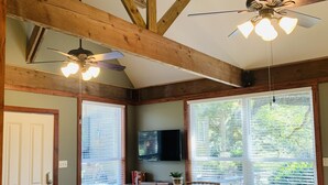 Amplia habitación con vigas de madera, ventilador de techo, televisión de pantalla plana y ventanas grandes con persianas.
