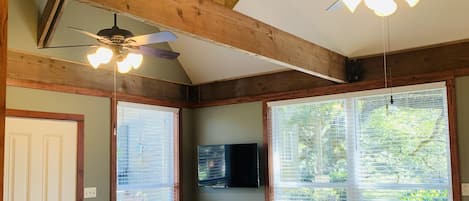 A spacious room with wooden beams, a ceiling fan, a flat-screen TV, and large windows with blinds.