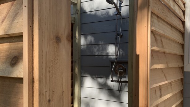 Una ducha exterior de madera con cabezal y manguera de metal, unida a una estructura de madera con revestimiento horizontal.