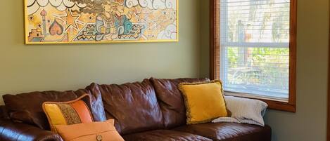A brown leather sofa with yellow and orange cushions, a wooden coffee table with a ceramic dish, and a colorful abstract painting on the wall.