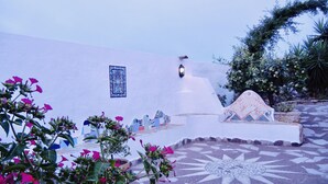 A courtyard with a tiled floor, a decorative wall with a blue tile, and a potted plant with pink flowers. - Alba B&B (Carloforte)