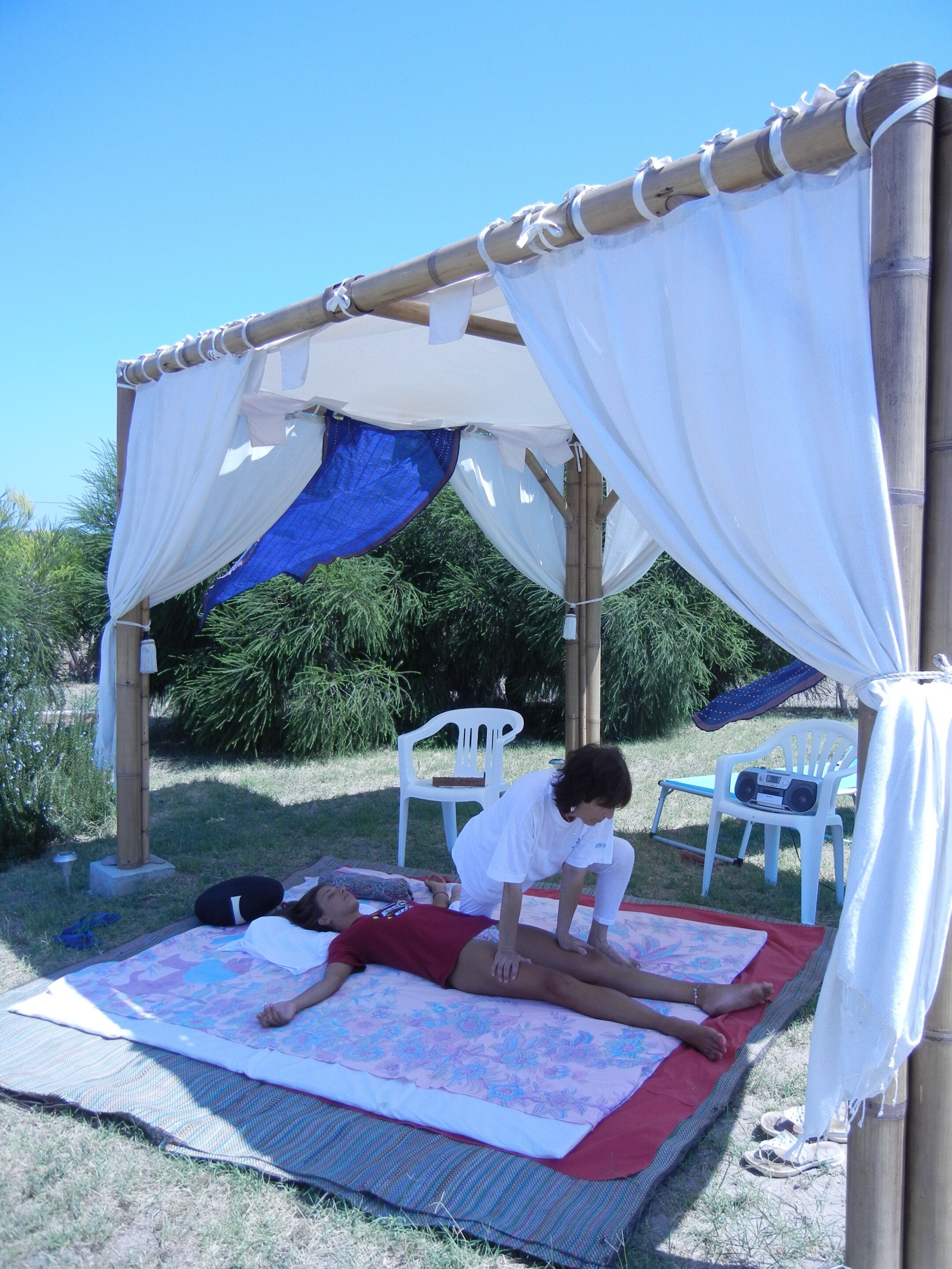 A covered outdoor area with a bamboo structure, two white plastic chairs, and a mat with a floral design.