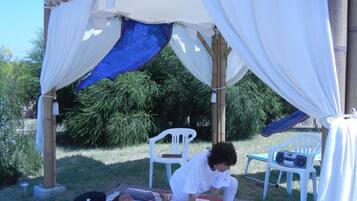 A covered outdoor area with a bamboo structure, two white plastic chairs, and a mat with a floral design.