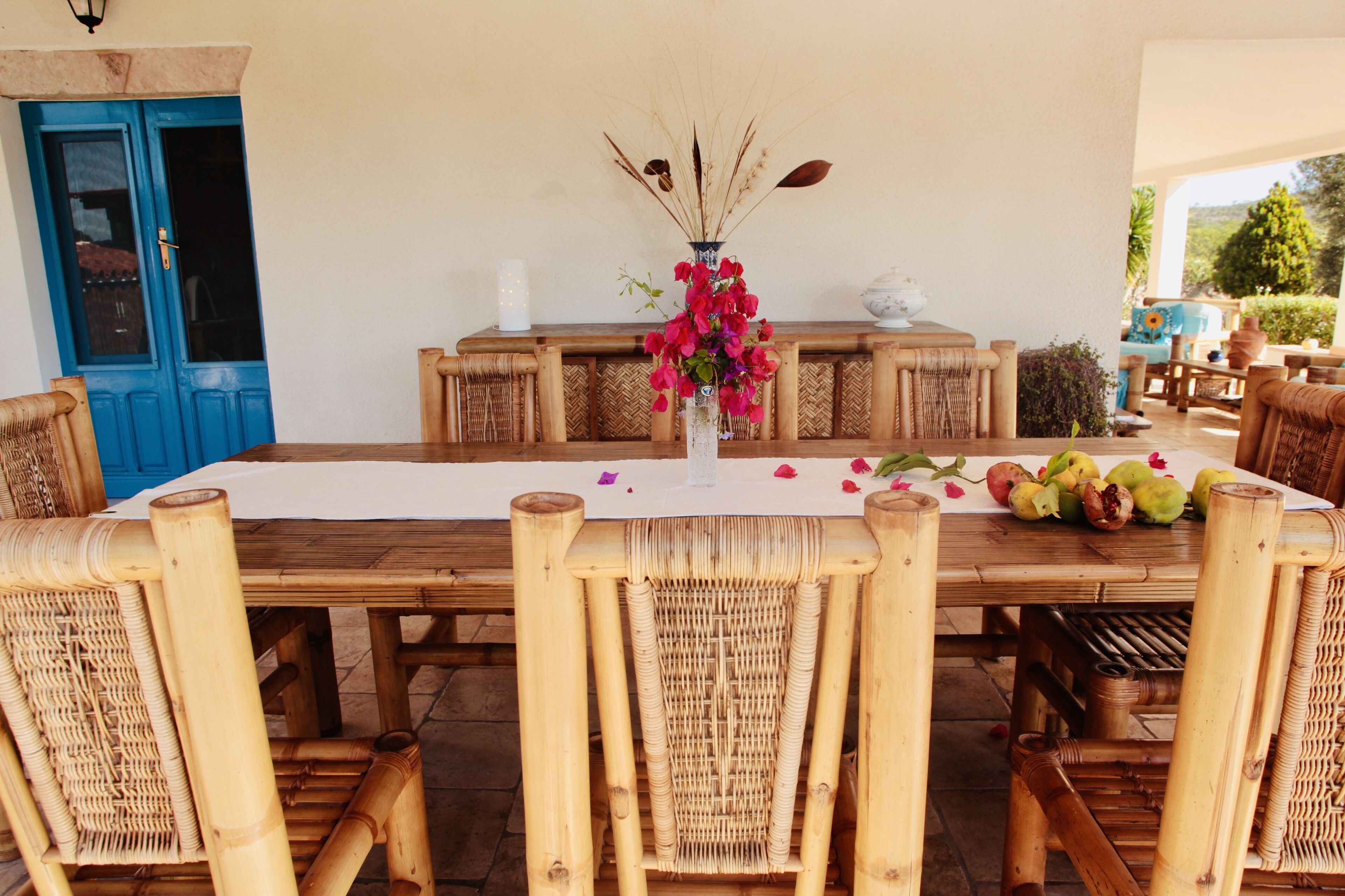 Uma mesa de jantar de madeira com toalha branca, rodeada por cadeiras de vime. Um vaso com flores cor-de-rosa e uma vela estão sobre a mesa.