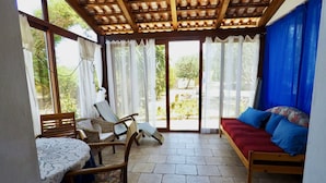 A covered outdoor seating area with a table and chairs, a sofa with cushions, and a view of greenery through large windows. - Alba B&B (Carloforte)