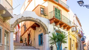 A narrow street with a stone archway leading to a building with a staircase, potted plants, and a bicycle parked outside. - Alba B&B (Carloforte)