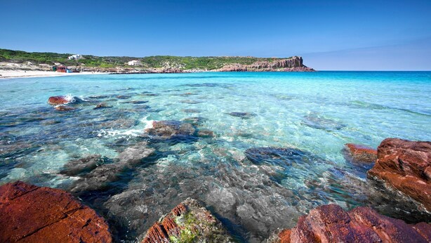 A coastal beach with clear turquoise water, rocky formations, and a distant cliff. - Alba B&B (Carloforte)