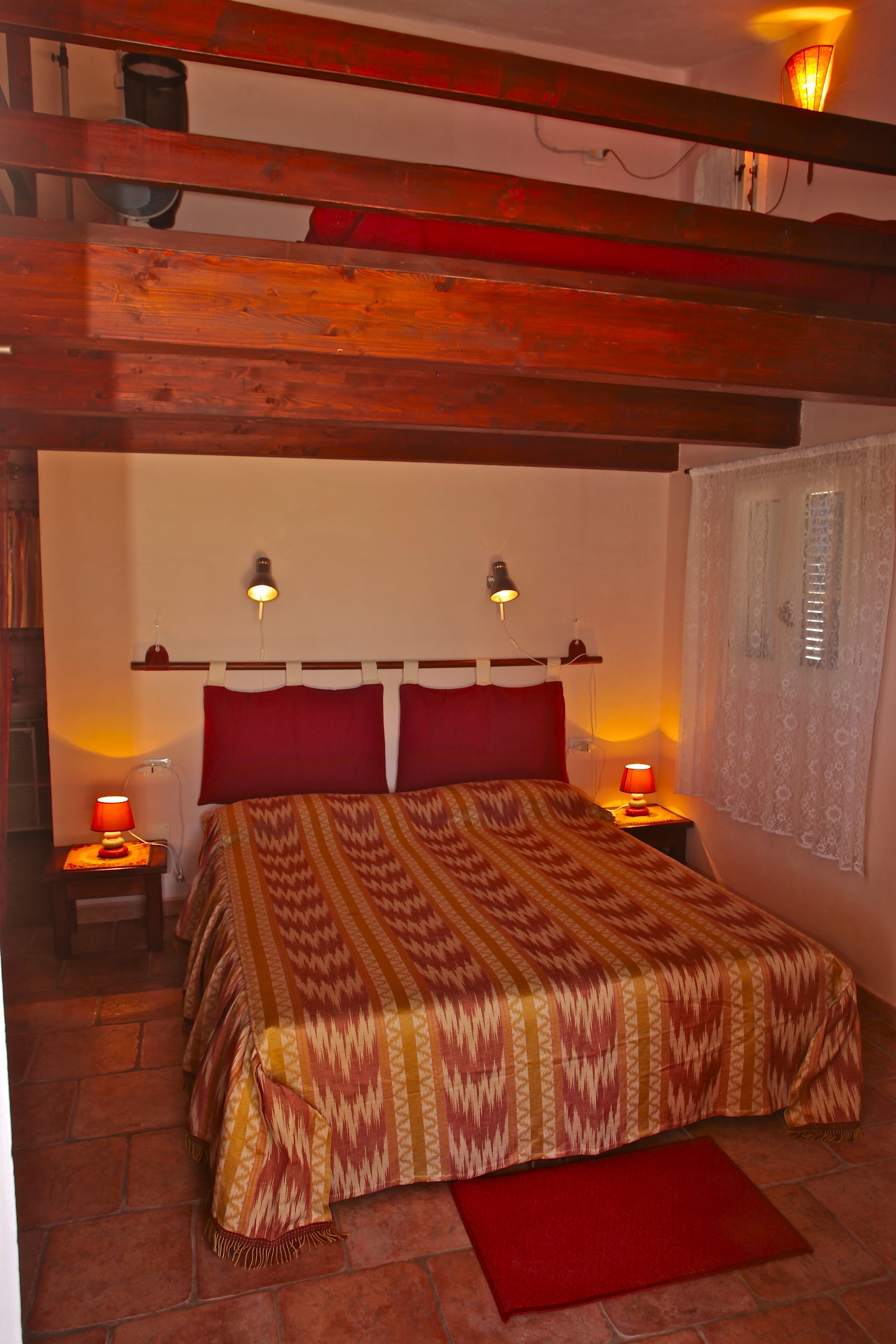 A bedroom with a bed, two bedside lamps, a red throw blanket, and a window with curtains.