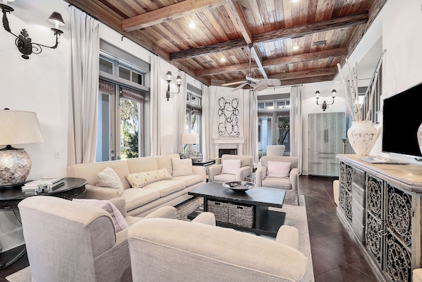 A spacious living room with a high ceiling, large windows, and a fireplace. There are multiple seating areas, including a sofa and chairs, and a coffee table.