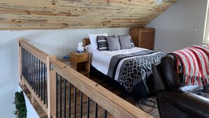 A bedroom with a sloped ceiling, wooden flooring, a bed with patterned bedding, a nightstand, a dresser, and a leather armchair. - Big Hole Valley cabin near river access (Wise River)