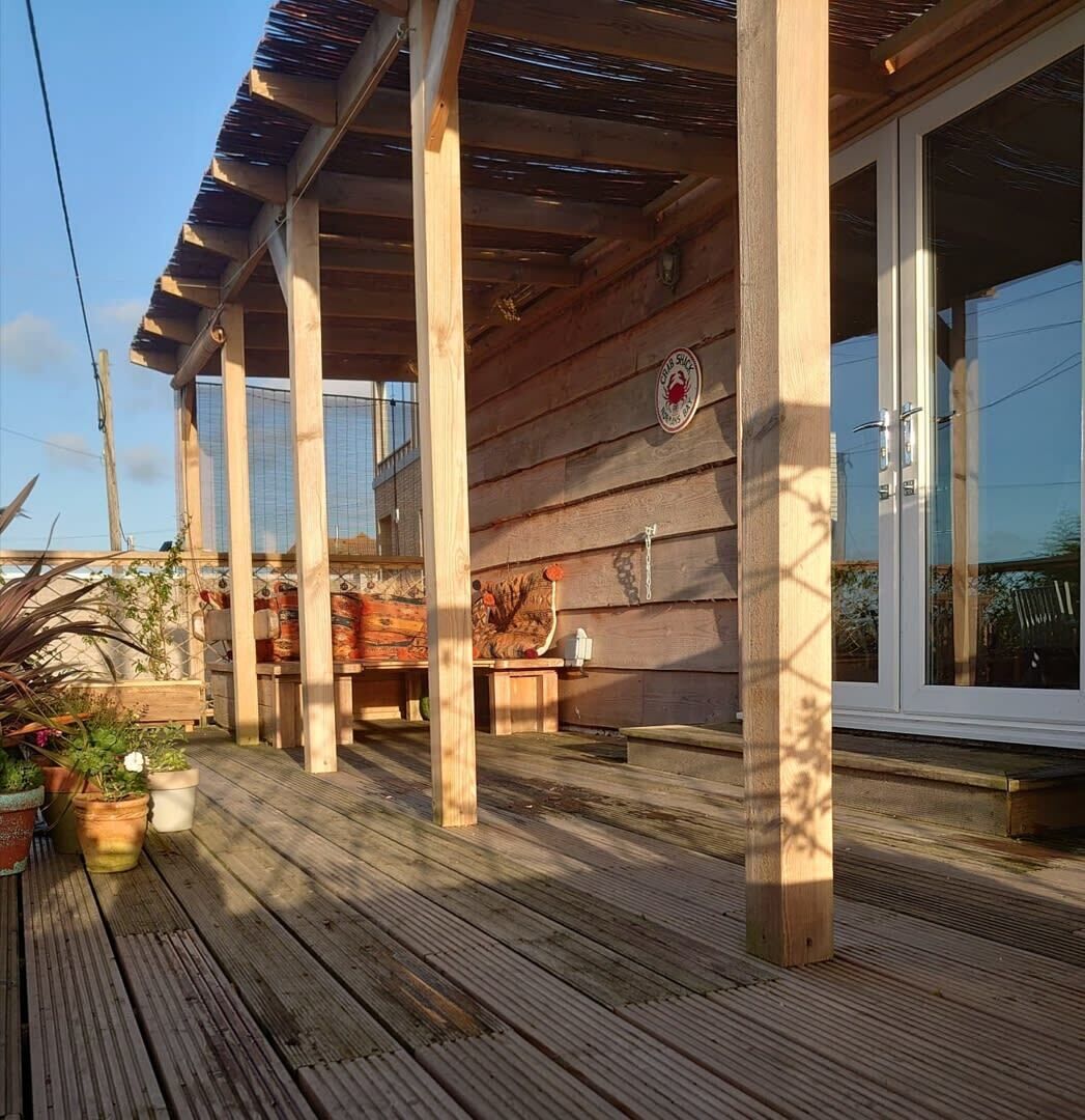 Une terrasse en bois dotée d’un espace salon abrité, de plantes en pot et d’une grande fenêtre.