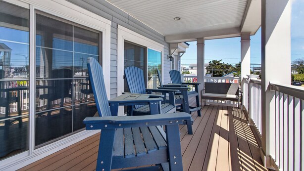 Una terraza techada al aire libre con sillas Adirondack azules y una mesa, ventanas grandes y una barandilla blanca.