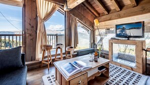L’intérieur d’un chalet en bois, avec une grande fenêtre, un canapé, une table avec un magazine et un téléviseur posé sur un meuble en bois.
