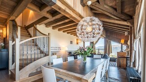 Un vaste espace dédié à la salle à manger, avec une table en bois et des chaises blanches, un escalier doté de rampes en bois et un grand luminaire décoratif.