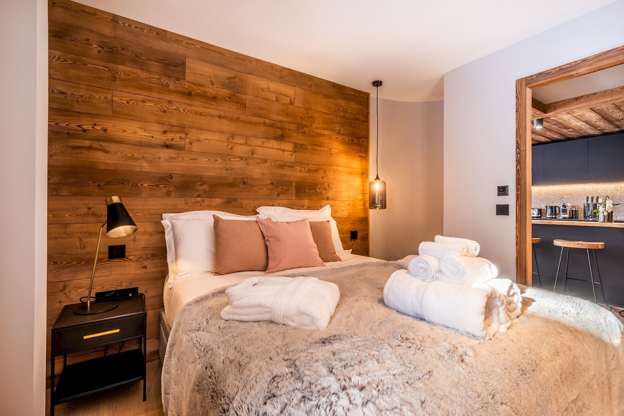 A bedroom with a wooden headboard, a bed with white linens, a fluffy rug, a bedside table with a lamp, and a bathroom visible through an open door.