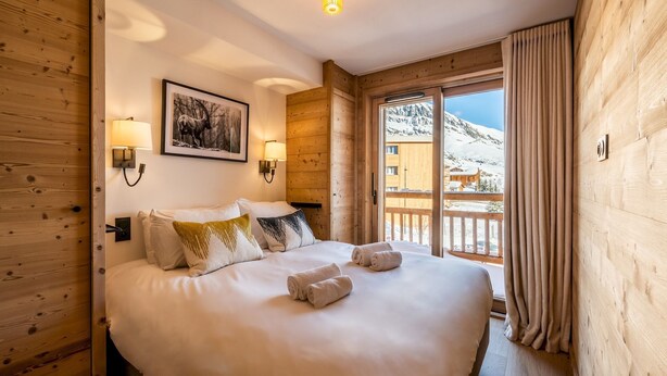 A wooden-paneled bedroom with a large bed, two wall-mounted lamps, a framed picture, and a view of a snowy mountain through a sliding door.