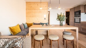 Una cocina moderna con armarios de madera, una isla central con taburetes, iluminación colgante y una zona de comedor con sofá y cojines.