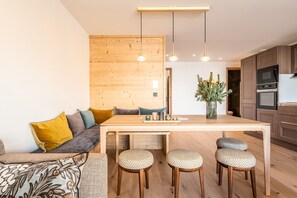 A modern kitchen with wooden cabinets, a central island with stools, pendant lighting, and a dining area with a sofa and cushions. - SIPO - Design apartment in brand-new residency (Huez)