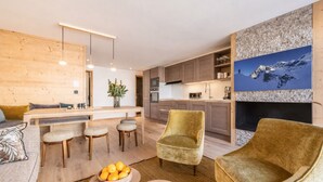 A modern living room with a sofa, armchairs, a coffee table, and a dining area with a table and chairs. There is a kitchen in the background with cabinets and a stove.