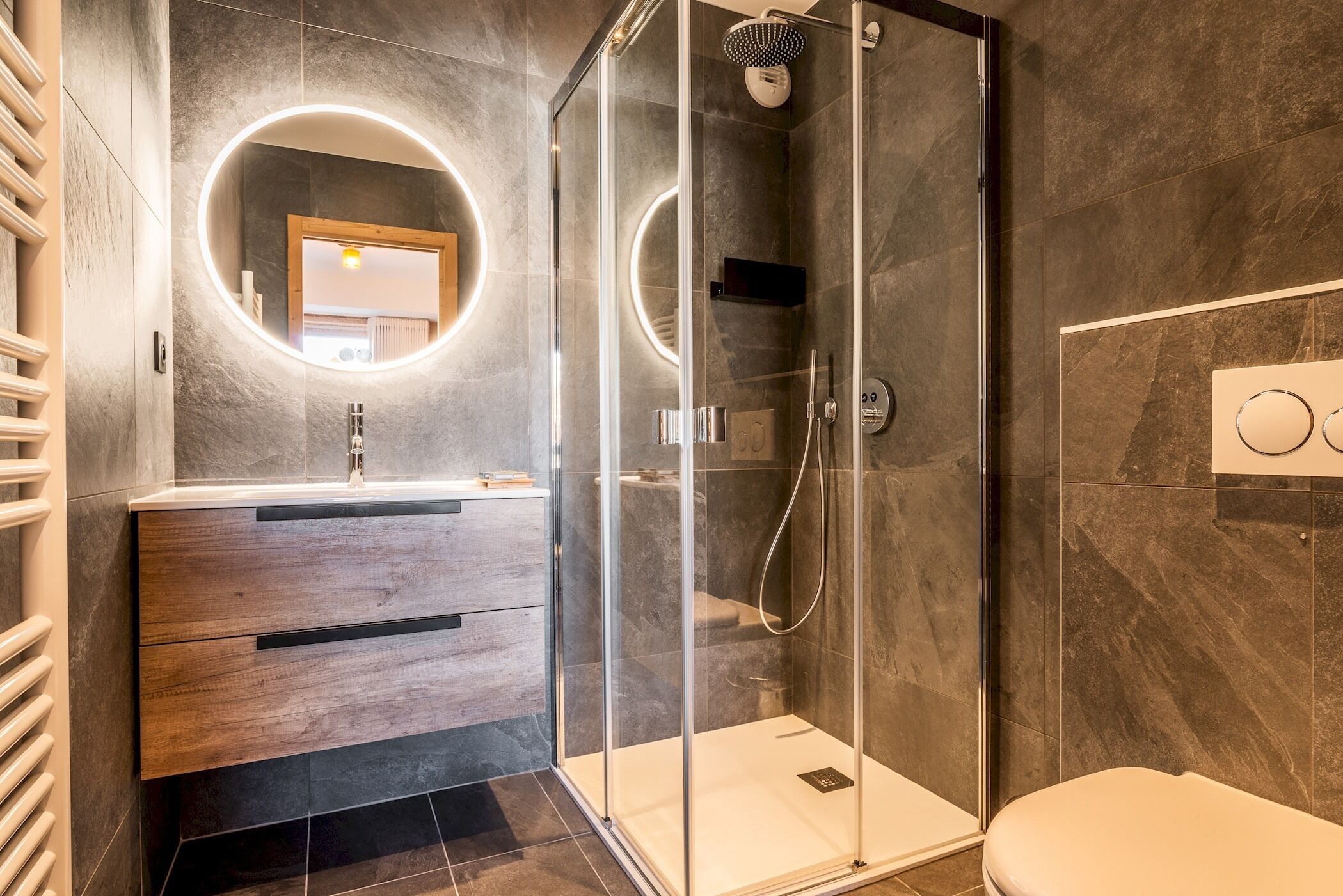 Een moderne badkamer met een douche met glazen wand, een houten wastafelmeubel met een ronde spiegel, en een toilet.