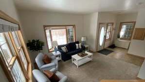 Amplia sala de estar con un sofá azul, un sillón a rayas y una mesa de centro blanca. Amplias ventanas ofrecen luz natural y vistas al exterior.