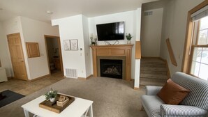 Una sala de estar con chimenea, un televisor montado encima, un sofá a rayas y una mesa de centro blanca con una bandeja.