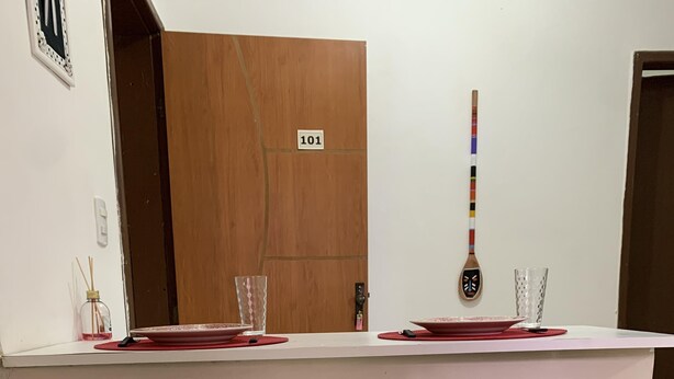A hotel room with a wooden door labeled '101', a white wall with a decorative mask, and a table set with red plates and glasses. - Oxe Rooms (Salvador)
