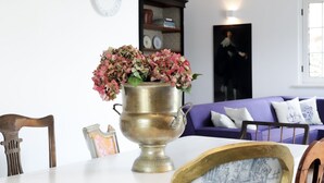 Een eettafel met een messing vaas vol roze bloemen, een lijst met foto, en een houten stoel. Op de achtergrond staat een paarse bank en een boekenkast.