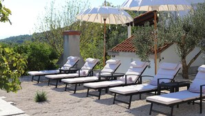 Een rij ligbedden met witte kussens en beige parasols, gelegen in een tuin met olijfbomen en een gebouw op de achtergrond.