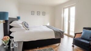 Een moderne slaapkamer met een groot bed, een nachtkastje met een bloevas en een fauteuil met kussens.