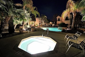 An outdoor swimming pool area with a hot tub, lounge chairs, and palm trees at night.