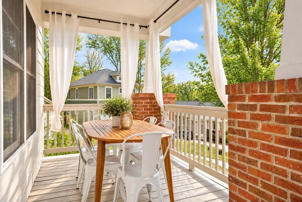 A covered outdoor patio with a wooden table and white chairs, a brick wall, and a view of a house in the background.