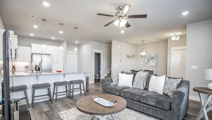A modern living room with a grey sofa, a coffee table, and a kitchen area with white cabinets and a stainless steel refrigerator. - Close to everything, New Fayetteville Home! (Fayetteville)
