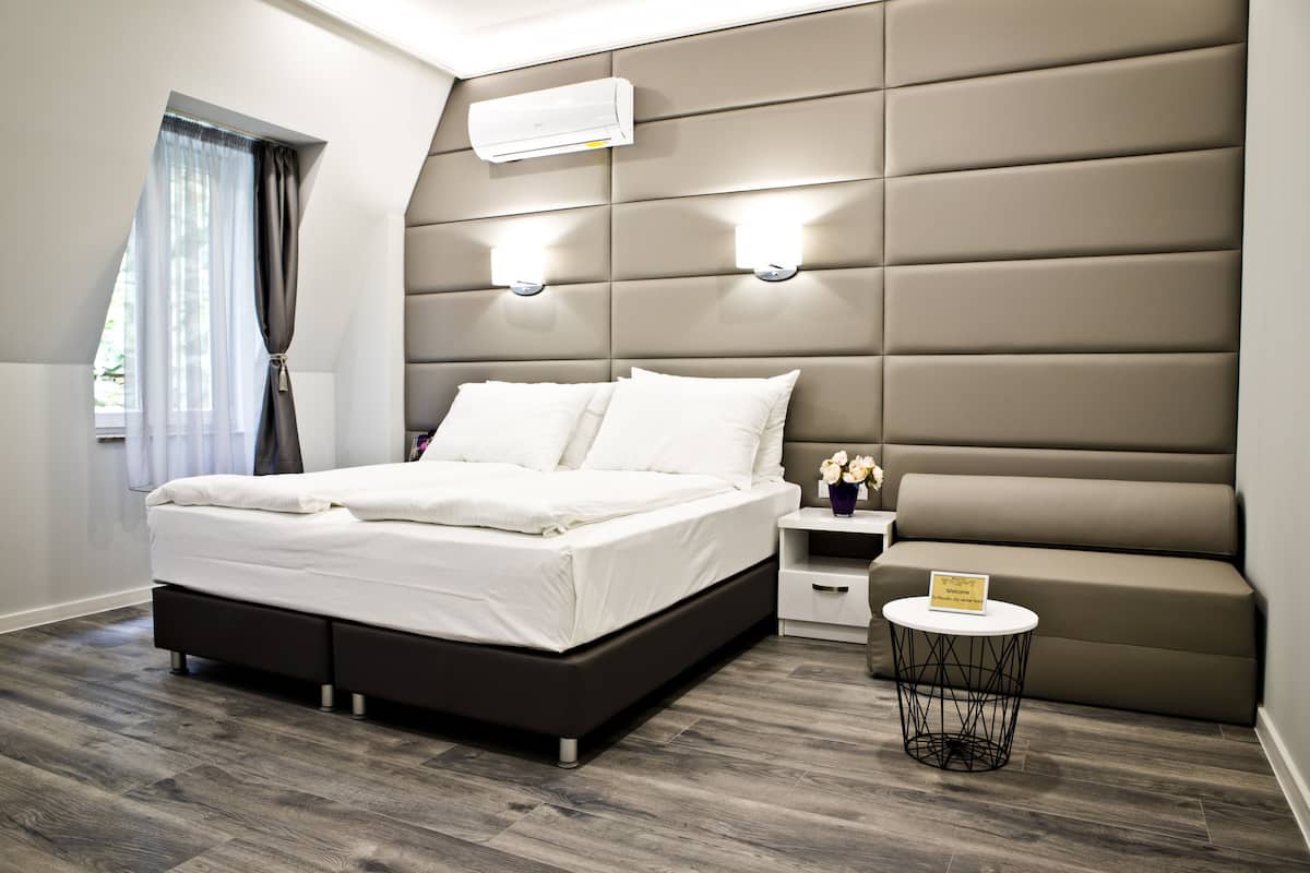 A modern bedroom with a large bed, a headboard with built-in lighting, a bedside table with a small lamp, and a window with curtains.