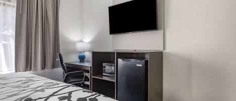 A hotel room with a bed, a desk with a computer, a TV, and a minibar.