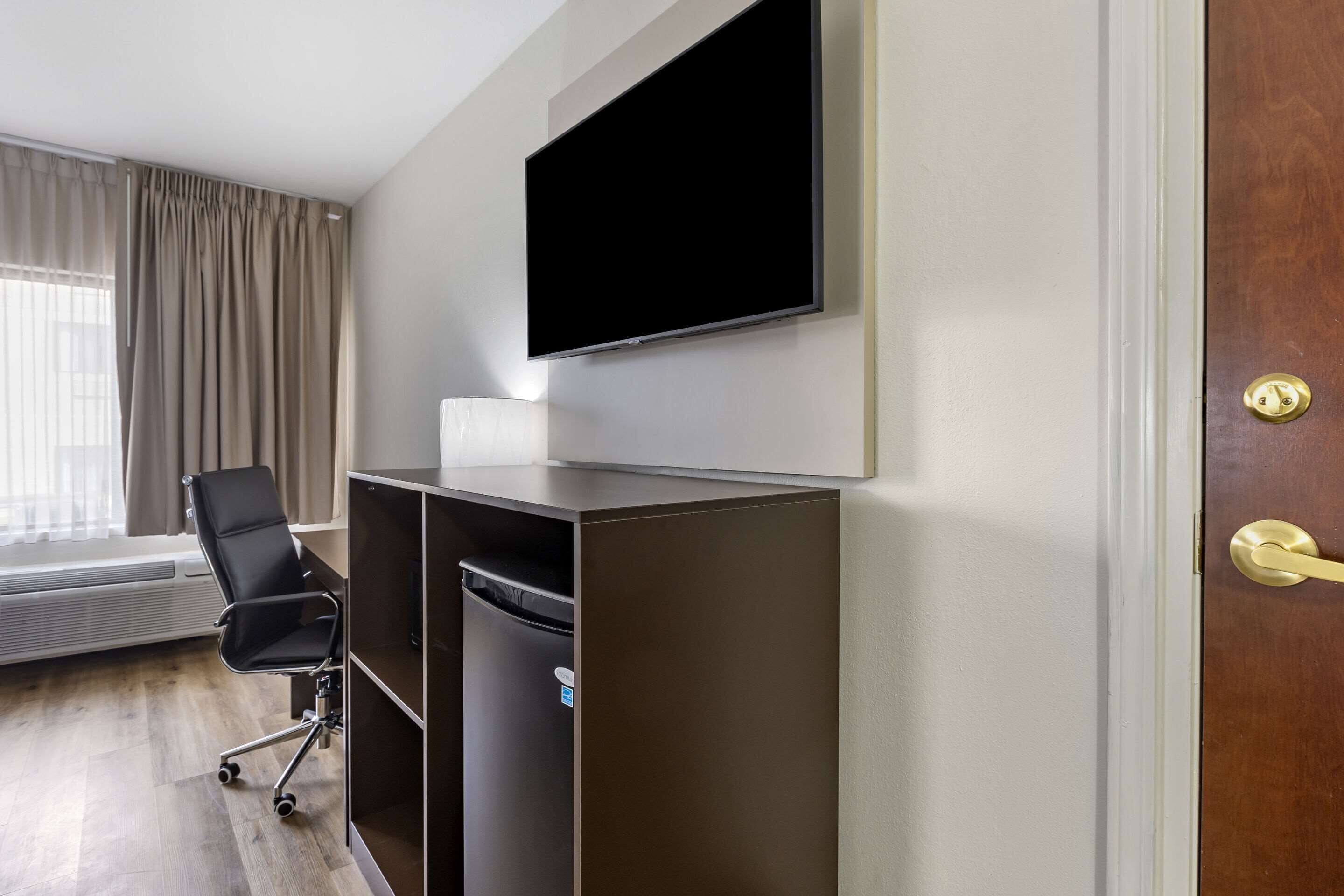 Une chambre d’hôtel équipée d’un téléviseur à écran plat fixé au mur, d’un fauteuil de bureau noir, d’une poubelle et d’une porte en bois avec une poignée en laiton.