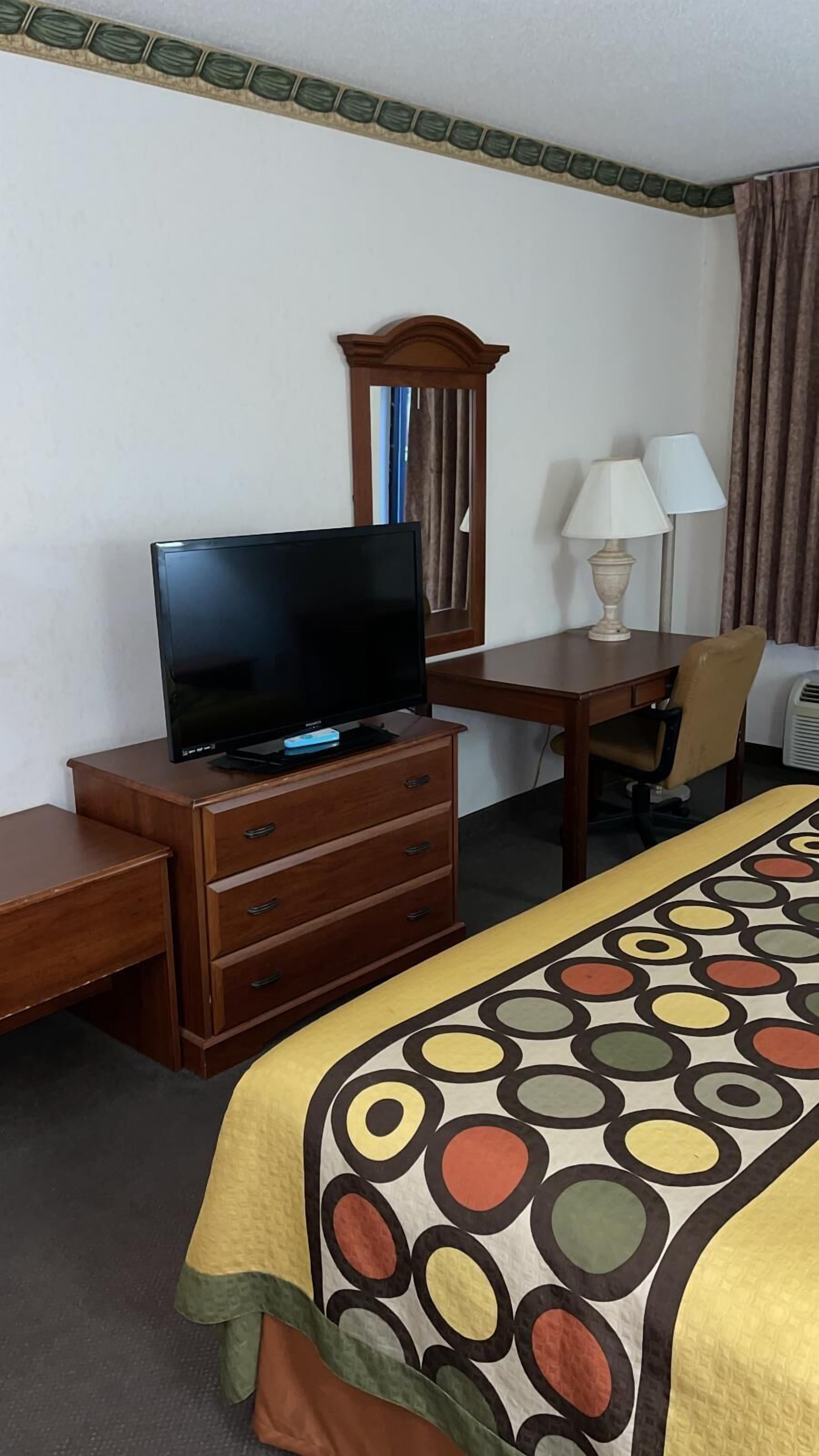 A hotel room with a bed, a desk with a television, a chair, a lamp, and a dresser.