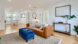 Amplia sala de estar con un sofá de cuero marrón, un reposapiés azul y un comedor con mesa y sillas blancas.