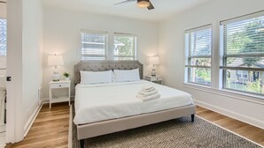 Un dormitorio ordenado con una cama grande, dos mesitas de noche con lámparas, un ventilador de techo y ventanas amplias con persianas.