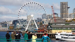 Blick auf das Wasser mit einem großen Riesenrad, einem in der Nähe liegenden Kreuzfahrtschiff und einer Skyline mit Gebäuden und einem Kran im Hintergrund.