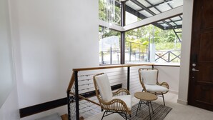 Una escalera con barandilla de madera, dos sillones de mimbre con cojines blancos, y vistas al verdor a través de amplias ventanas.
