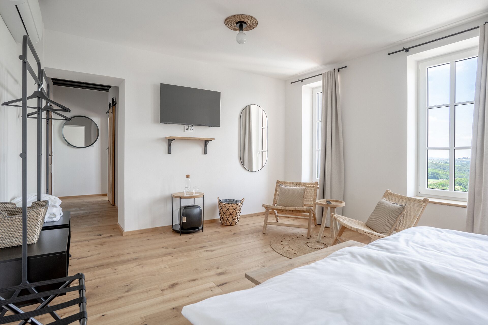 Una habitación de hotel moderna con una cama grande, un televisor montado en la pared, una silla de mimbre y vistas al exterior a través de la ventana.