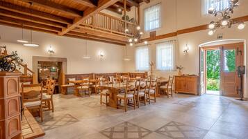 A spacious dining area with wooden tables and chairs, a high ceiling with exposed beams, and a view of the outdoors through large windows.