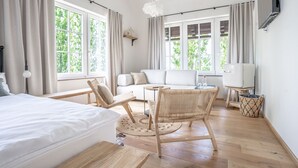 A modern hotel room with a bed, a sofa, a dining table, wicker chairs, and large windows offering a view of greenery. - SIBON Wine & Spa Resort Jeruzalem (Ljutomer)