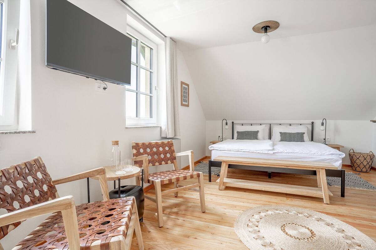 Un dormitorio con cama, dos sillas, una mesita, una ventana y una lámpara en el techo.
