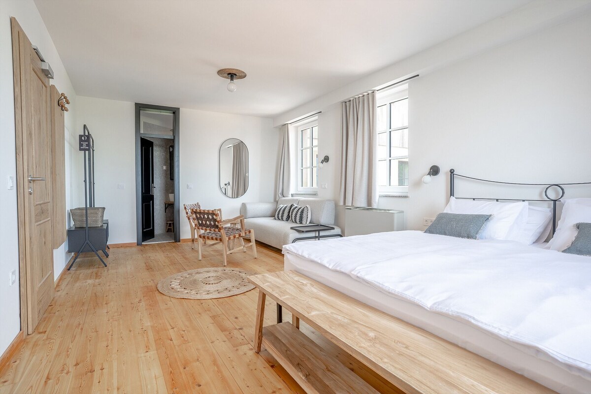 Une chambre avec un plancher en bois, un lit, un banc, une chaise, un miroir et une porte.