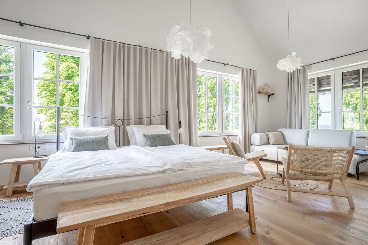 Un dormitorio con cama, banca de madera, silla de mimbre y ventanas amplias.