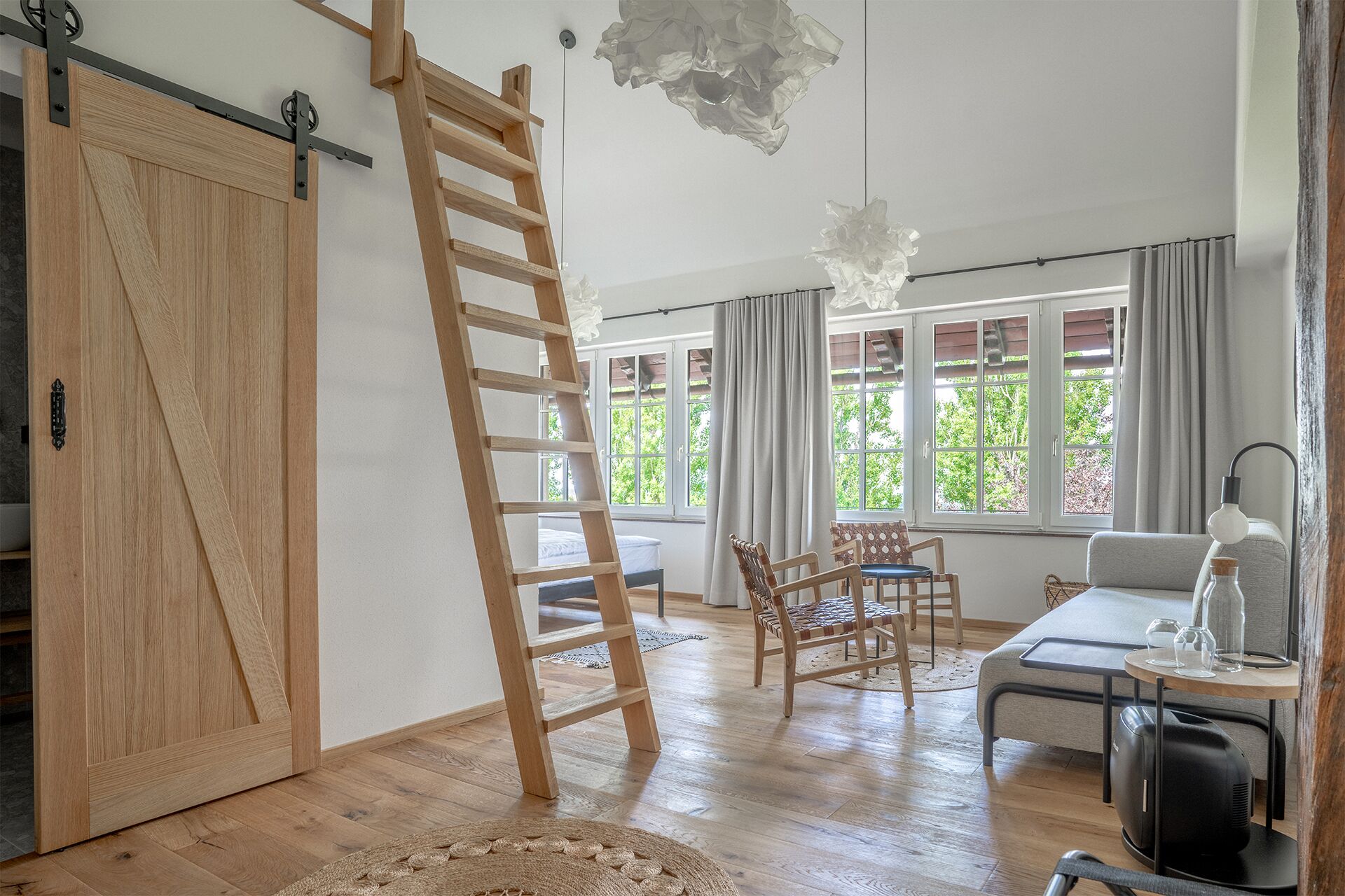 A spacious room with a wooden floor, a sliding door, a wooden ladder, a dining area with chairs and a table, and large windows with curtains.