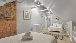 A bedroom with a brick wall, a bed with white bedding, a bedside table, a chair, and a small desk with a chair.